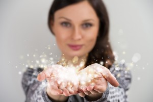 Pretty young brunette woman smiling against gray background with magic sparkle in her hands cupped together